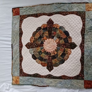 May include: A square quilt with a white background and a brown and green circular design in the center. The quilt is surrounded by a blue and brown border.