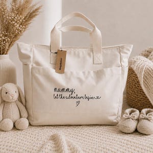 May include: A cream-coloured canvas tote bag featuring two top handles and a front pocket. The text "mummy, let the adventure begin" is printed in black script on the front, accompanied by a small heart icon. A brown cardboard tag is attached.
