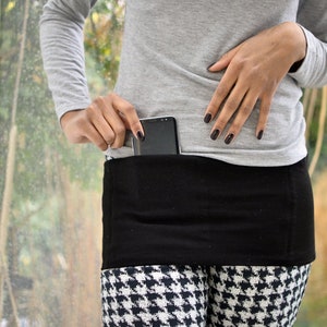 Puede incluir: Un cinturón de tela negro con un bolsillo para un teléfono inteligente. El cinturón se lleva sobre leggings negros y blancos de pata de gallo.