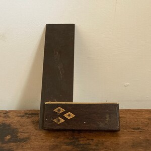 May include: A vintage wooden and metal try square with brass accents and four diamond-shaped inlays. The try square is resting on a wooden surface.