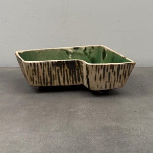 Puede incluir: Una jardinera rectangular de cerámica con una forma única. El exterior tiene un patrón texturizado, similar a la corteza, en beige y marrón. El interior es de un verde vibrante. La jardinera está diseñada con un recorte distintivo en un lado.