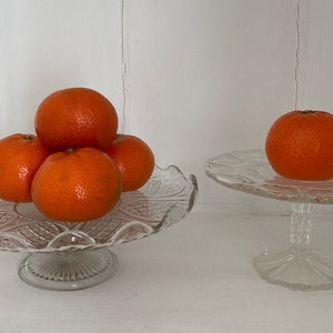May include: Two glass cake stands with a stack of four orange mandarins on the larger stand and a single mandarin on the smaller stand.