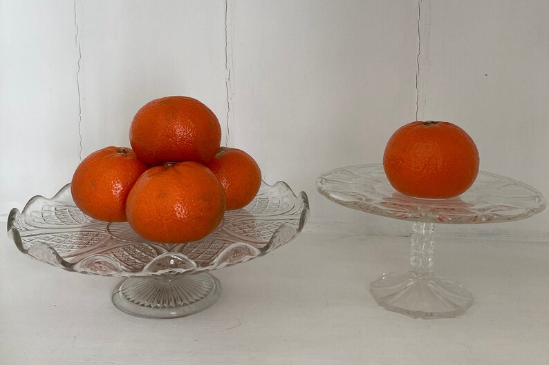 May include: Two glass cake stands with a stack of four orange mandarins on the larger stand and a single mandarin on the smaller stand.
