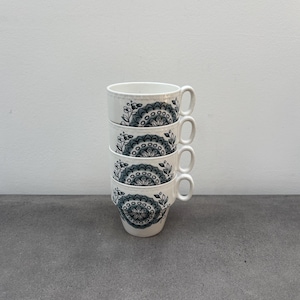 Könnte beinhalten: Vier weiße Keramikbecher gestapelt, jeder mit Henkel und einem dekorativen floralen und geometrischen Muster in Teal und Grau. Die Becher stehen auf einer grauen Oberfläche vor einem weißen Hintergrund.