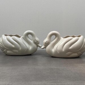 May include: Two cream-coloured ceramic swan planters. Each swan has a curved neck and a ruffled edge along the top. The swans are facing each other, with their beaks almost touching. The planters are on a grey surface.