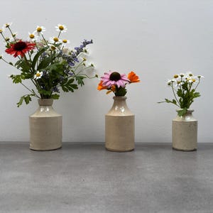 May include: Three cylindrical cream-coloured ceramic bottles of varying heights, each functioning as a vase. The largest holds a bouquet of white daisies and red flowers, the middle holds purple and orange flowers, and the smallest holds white daisies.