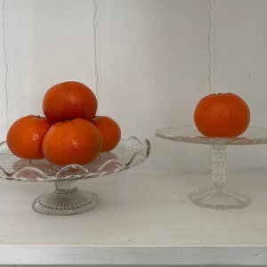 May include: Two glass cake stands with a stack of orange mandarins on one stand and a single mandarin on the other.