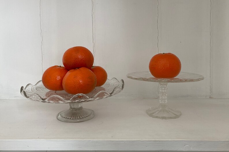 May include: Two glass cake stands with a stack of orange mandarins on one stand and a single mandarin on the other.