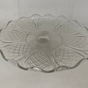 May include: A clear glass cake stand with a scalloped edge and a decorative pattern of diamond shapes. The stand has a raised center and a smooth, flat surface.