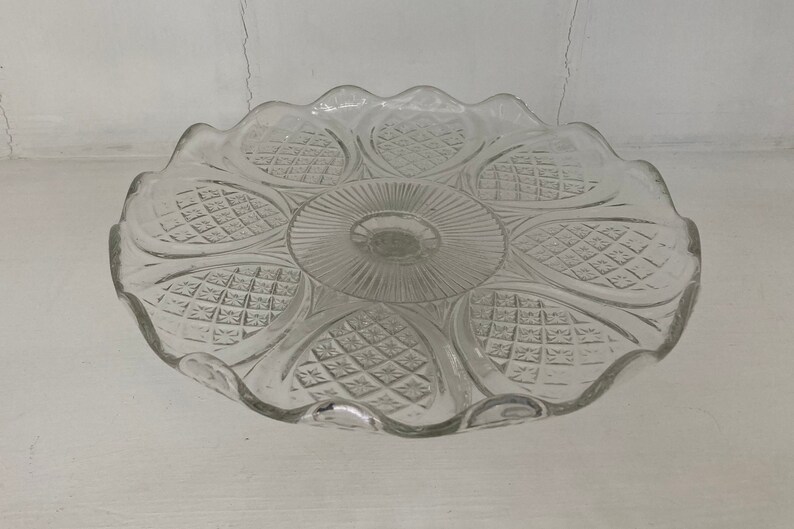 May include: A clear glass cake stand with a scalloped edge and a decorative pattern of diamond shapes. The stand has a raised center and a smooth, flat surface.