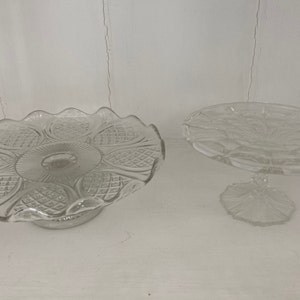 May include: Two clear glass cake stands with intricate patterns. The larger stand has a scalloped edge and a wider base. The smaller stand has a round base and a more delicate design.