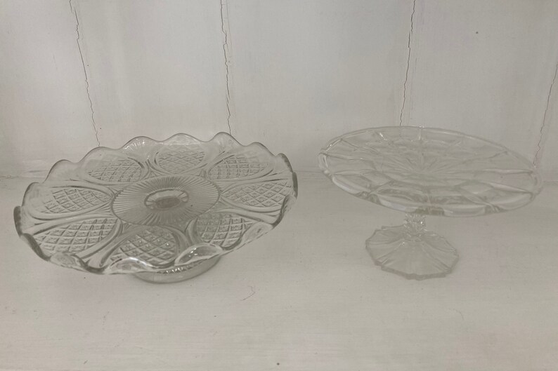 May include: Two clear glass cake stands with intricate patterns. The larger stand has a scalloped edge and a wider base. The smaller stand has a round base and a more delicate design.