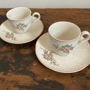 May include: Two white teacups and saucers with a floral pattern. The cups have a pink rim and the saucers have a delicate lace pattern.