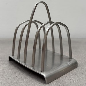 May include: A silver-toned metal toast rack featuring a rectangular base and five arched dividers of varying heights. The structure is composed of smooth, thin metal bars arranged to create four distinct slots for holding slices of toast upright.