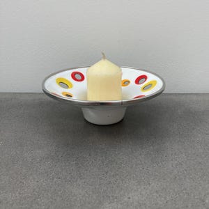 May include: White ceramic candle holder with a silver rim and a red and yellow polka dot design. A white candle is placed in the holder.