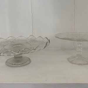 May include: Two clear glass cake stands with scalloped edges. The larger stand has a more intricate design and a wider base. The smaller stand has a simpler design and a narrower base.