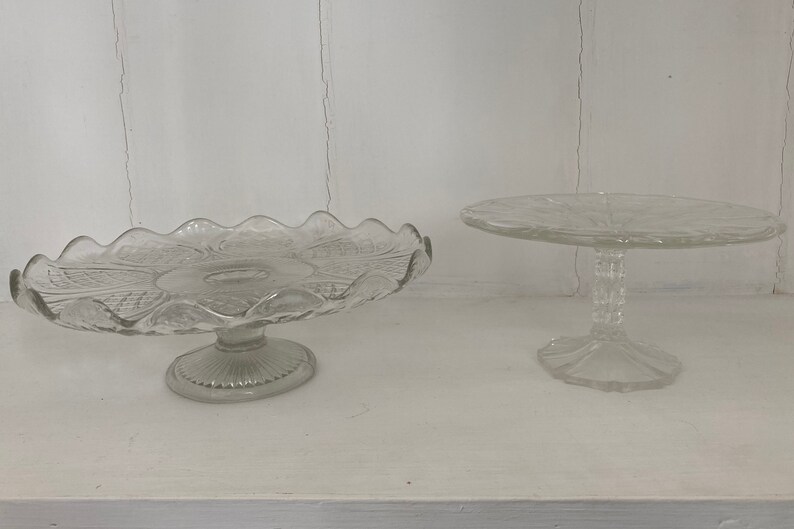 May include: Two clear glass cake stands with scalloped edges. The larger stand has a more intricate design and a wider base. The smaller stand has a simpler design and a narrower base.