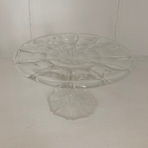 May include: A clear glass cake stand with a scalloped edge and a pedestal base. The stand is designed with a delicate, intricate pattern.