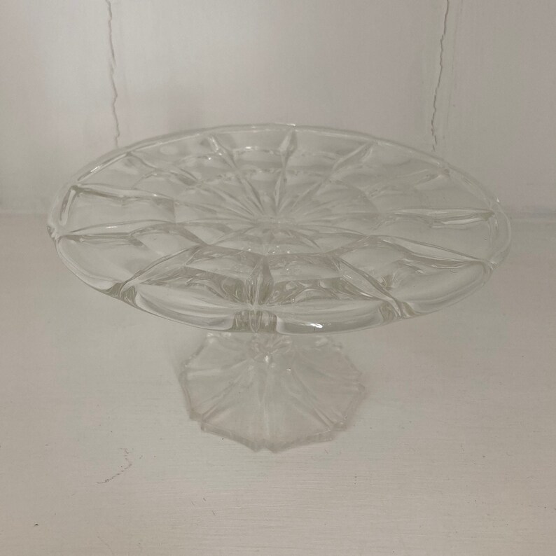 May include: A clear glass cake stand with a scalloped edge and a pedestal base. The stand is designed with a delicate, intricate pattern.