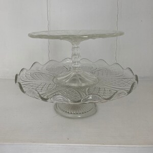 May include: A two-tiered glass cake stand with a clear, cut-glass design. The stand has a scalloped edge and a pedestal base.