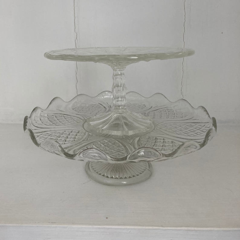 May include: A two-tiered glass cake stand with a clear, cut-glass design. The stand has a scalloped edge and a pedestal base.