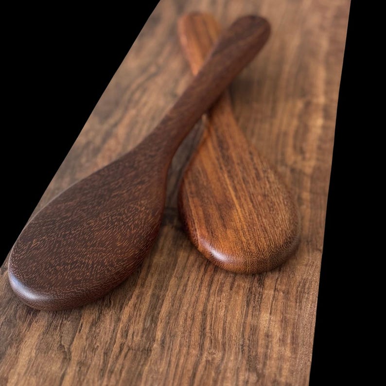 May include: Two wooden spoons made of dark brown wood. The spoons are crossed over each other and are lying on a wooden surface.