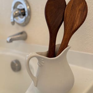 May include: Two wooden spoons are placed in a white ceramic jug with a handle. The jug is sitting on a white countertop.
