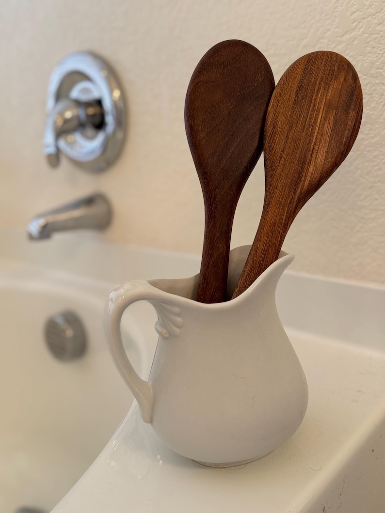 May include: Two wooden spoons are placed in a white ceramic jug with a handle. The jug is sitting on a white countertop.