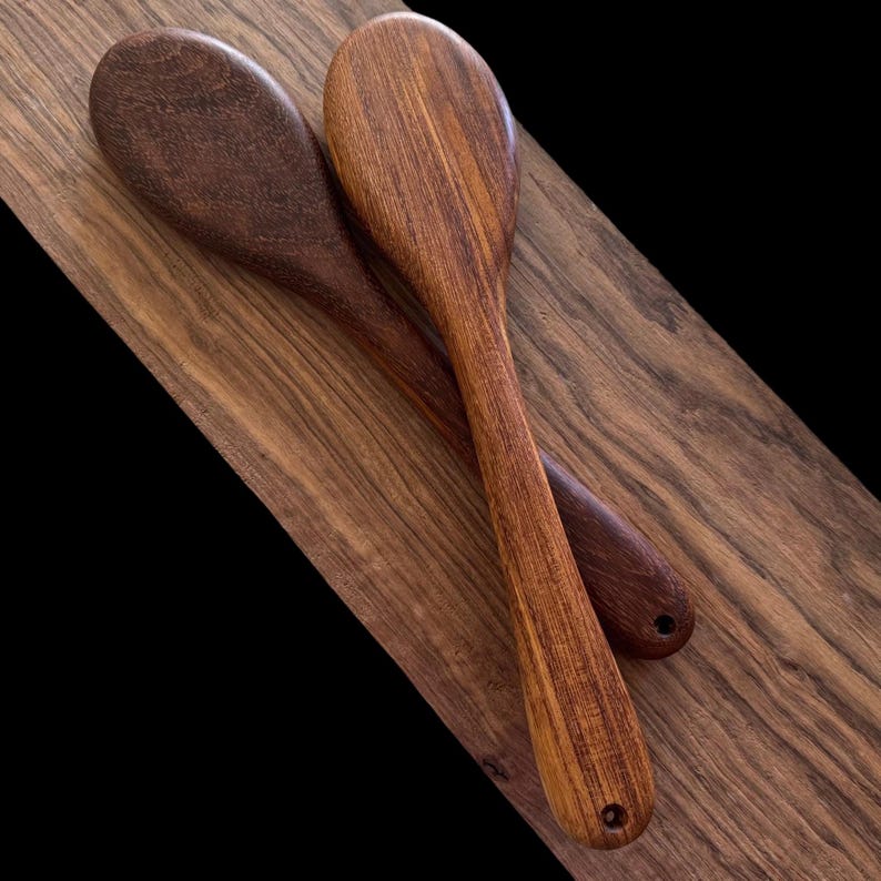 May include: Two wooden spoons, one brown and one reddish-brown, are lying on a wooden surface. The spoons are made of different types of wood, and the grain patterns are visible.