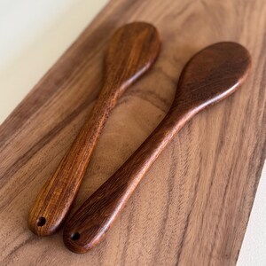 May include: Two wooden spoons made of dark brown wood. The spoons are lying on a wooden surface.