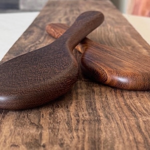May include: Two wooden spoons made of dark brown wood. The spoons are oval shaped and have a smooth, polished finish.