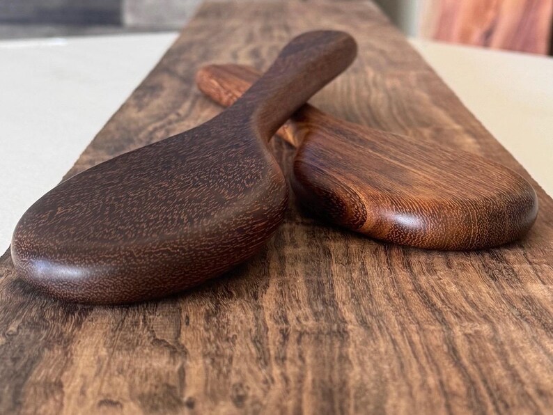 May include: Two wooden spoons made of dark brown wood. The spoons are oval shaped and have a smooth, polished finish.