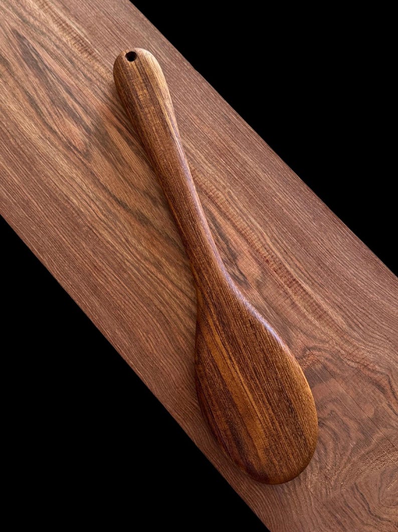 May include: A wooden spoon with a dark brown colour and a natural wood grain pattern. The spoon has a long handle and a rounded bowl.