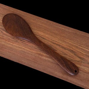 May include: A dark brown wooden spoon with a rounded bowl and a long handle. The spoon is lying on a wooden surface.