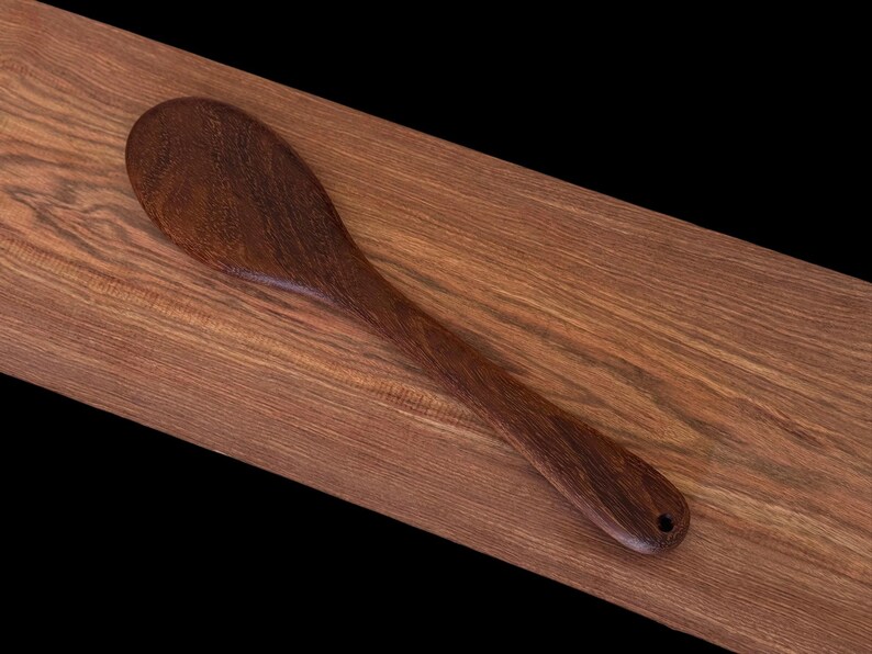 May include: A dark brown wooden spoon with a rounded bowl and a long handle. The spoon is lying on a wooden surface.