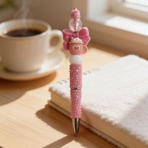 May include: A pink ballpoint pen featuring a barrel covered in small pink rhinestones. The top of the pen is decorated with a white bead, a pink bow, a miniature pink mug charm with a white heart, a clear faceted bead, and a pink polka-dot bead.