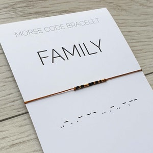 Puede incluir: Un brazalete de cuerda marrón con cuentas negras y doradas que deletrean la palabra "FAMILY" en código Morse. El brazalete está sobre una tarjeta blanca con las palabras "Morse Code Bracelet" y "FAMILY" impresas en ella. La tarjeta también incluye el código Morse para la palabra "FAMILY" y el logotipo "MC-bracelets".