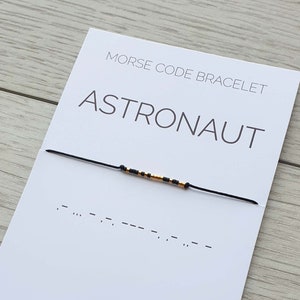 Pode incluir: Um bracelete de corda preto com contas douradas que soletram "ASTRONAUT" em código Morse. O bracelete está em um cartão branco com o texto "MORSE CODE BRACELET" e "ASTRONAUT" em texto preto. O cartão também tem o texto ".MC-bracelets" em texto preto.