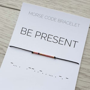 May include: A white card with black text that reads "Morse Code Bracelet" and "BE PRESENT". The card also includes a black string bracelet with pink and gold beads. The bracelet is tied in a knot and the morse code for "BE PRESENT" is written below the bracelet.