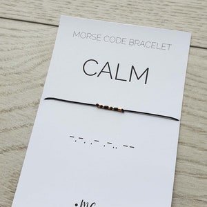 May include: A black string bracelet with gold beads spelling out the word "CALM" in Morse code. The bracelet is on a white card with the words "Morse Code Bracelet" and "CALM" printed on it. The card also has the text ".MC-bracelets" printed on it.