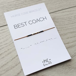 Puede incluir: Una tarjeta blanca con el texto "Morse Code Bracelet" y "BEST COACH" en negro. Un brazalete de cuerda marrón con cuentas negras se muestra en la tarjeta. El código Morse para "BEST COACH" está escrito debajo del brazalete.