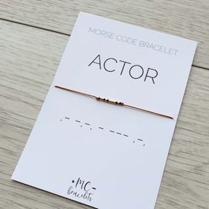 May include: A white card with the text "MORSE CODE BRACELET" and "ACTOR" printed on it. A brown string bracelet with black beads is on the card. The morse code for "ACTOR" is printed below the bracelet. The text ".MC-bracelets" is printed in the bottom right corner of the card.
