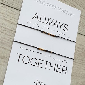 Pode incluir: Duas pulseiras de corda preta com contas douradas que soletram as palavras "ALWAYS" e "TOGETHER" em código Morse em um cartão branco com o texto "MORSE CODE BRACELET" e ".MC-bracelets".