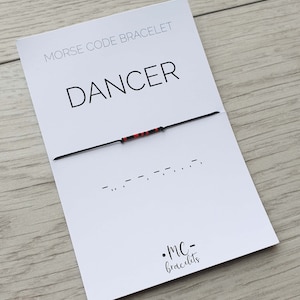 May include: A black string bracelet with red beads spelling out the word "DANCER" in Morse code. The bracelet is on a white card with the words "Morse Code Bracelet" and "DANCER" in black text. The card also has the text ".MC-bracelets" in black text.
