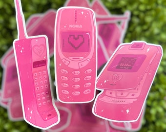 May include: Three pink cell phone stickers with a heart design. The phones are a flip phone, a Nokia phone, and a basic cell phone.