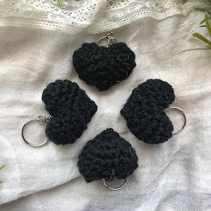 May include: Four black crocheted heart-shaped keychains with silver key rings. The keychains are displayed on a white fabric background.