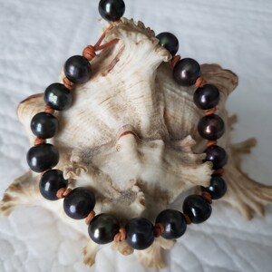 May include: A black pearl bracelet with brown leather cord. The bracelet is on a white seashell.