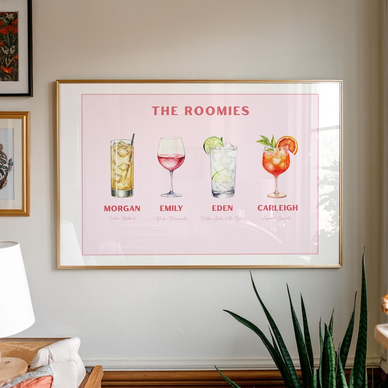 May include: A framed print with four cocktail illustrations. The print is titled "THE ROOMIES" and features the names Morgan, Emily, Eden, and Carleigh. The background is a pale pink. The cocktails are in various glasses.