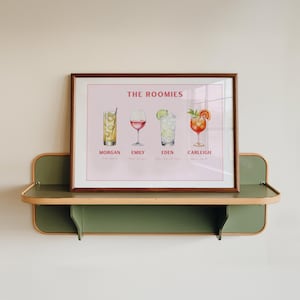 May include: A framed print displaying four watercolour cocktail illustrations. The print, titled "THE ROOMIES," includes drink names and individual names: Morgan, Emily, Eden, and Carleigh. The artwork is displayed on a green and wood-toned shelf.