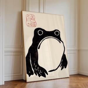 May include: A framed art print featuring a minimalist black frog illustration on a cream background. The frog has large eyes and a wide mouth. A red stamp-like symbol is in the upper left corner. The frame is gold.